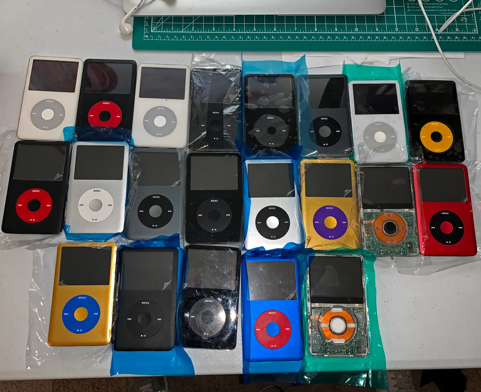An overhead shot of two dozen iPod Classics arranged in rows on a cutting mat — different colors, different generations, all stripped down for modding.